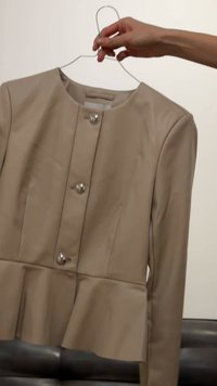 Χέρι που κρατά μια μπεζ ζακέτα με κουμπιά σε συρμάτινη κρεμάστρα, μπροστά σε απλό φόντο.