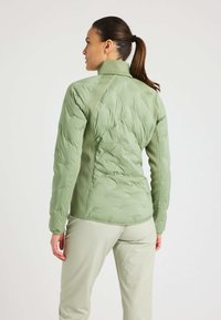 Chaqueta verde aislante con textura acolchada y cuello alto, combinada con pantalones a cuadros. Enfócate en los detalles del material y la costura.
