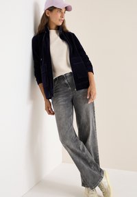 Chaqueta de corduroy navy con cremallera y dos bolsillos frontales, suéter de punto crema, jeans de pierna ancha gris y zapatillas amarillas claras. Una gorra rosa completa el look.