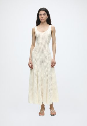 Frau steht und trägt ein ärmelloses weißes gehäkeltes Maxikleid und beige Riemchensandalen vor einem schlichten hellgrauen Hintergrund.