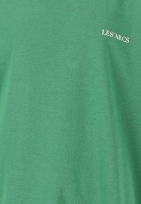 Grøn bomulds T-shirt med en blød tekstur og hvid trykt tekst, der læser "LES'ARCS" på den øverste venstre side.