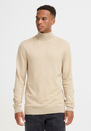 Jersey de cuello alto en color beige, confeccionado en tejido de punto acanalado, con mangas largas y un diseño ajustado. Textura suave y un leve estiramiento.