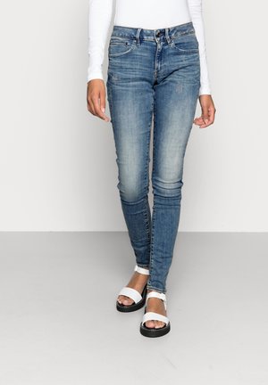 Un jean skinny en denim bleu clair avec une finition délavée. Présente des poches avant et une fermeture à boutons classique. Associé à des sandales blanches.