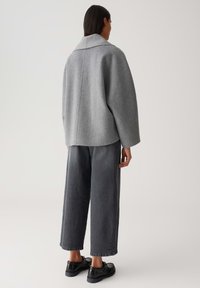 Vrouw in een losse lichtgrijze jack, met brede donkergrijze jeans en zwarte schoenen, staand met haar rug naar de camera.