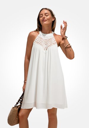Mujer con vestido blanco sin mangas con escote de crochet, sosteniendo un bolso tejido, con los ojos cerrados y levantando una mano cerca de su rostro.
