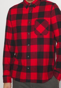 Camisa de cuadros en rojo y negro, de manga larga, con bolsillo frontal y dobladillo redondeado. Presenta botones negros y una tela suave y texturizada.