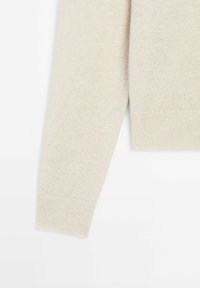Manche longue de pull beige tricoté avec un poignet côtelé sur fond blanc.
