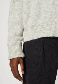 Šviesiai pilkas megztinis su raštuotu apvadu ir rankogaliais, pasižymintis švelnia tekstūra. Matosi tamsios kelnės po juo. Ranką padėti ant rankovės.