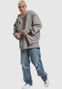 Gray hoodie with front zip and large pockets, layered over a gray t-shirt. Distressed blue denim jeans and white sneakers complete the outfit.