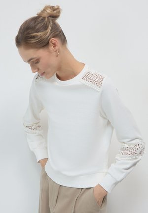 Donna con i capelli raccolti in un bun, indossa un top bianco a maniche lunghe con dettagli in pizzo sulle spalle e sulle maniche, e pantaloni beige con le mani nelle tasche.