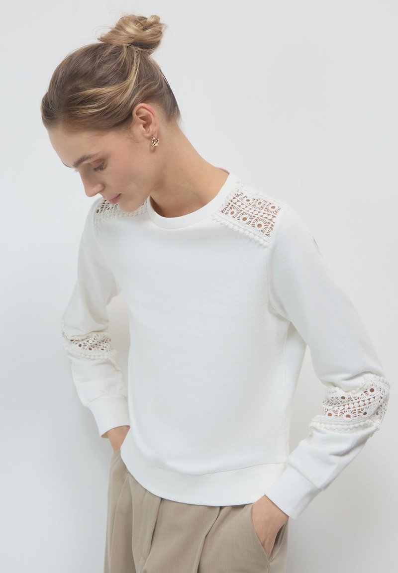 Donna con i capelli raccolti in un bun, indossa un top bianco a maniche lunghe con dettagli in pizzo sulle spalle e sulle maniche, e pantaloni beige con le mani nelle tasche.