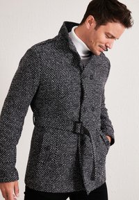 Cappotto intrecciato in bianco e nero con finitura testurizzata, caratterizzato da un colletto alto, doppi bottoni e un dettaglio a cintura in vita.