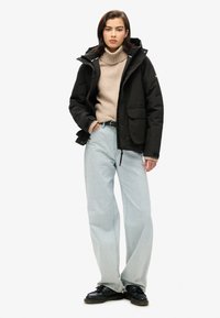 Zwarte waterdichte jas met capuchon over een beige coltrui, gecombineerd met lichtblauwe wijde jeans en zwarte schoenen. Kenmerken zijn onder andere voorzakken.