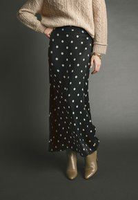 Zwarte satijnen rok met witte stippen, veel langer dan de vloer, in een vloeiende uitstraling. Gecombineerd met beige haklaarzen en een crèmekleurige gebreide trui.