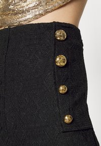 Svarta strukturerade högmidjade byxor med gulddekorativa knappar på sidan, med ett detaljerat mönster som liknar spets.
