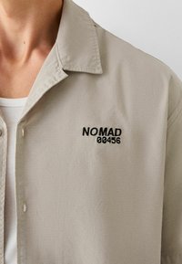 Camisa de botones de un beige claro con un acabado texturizado, que presenta un texto bordado en negro "NO MAD 00456" en el lado izquierdo del pecho.