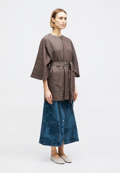 Γυναίκα που φοράει χαλαρό καφέ παλτό με ζώνη και φαρδιά μανίκια, μια μπλε τζιν φούστα με κουμπιά και μπεζ μπαλαρίνες, στέκεται μπροστά σε μια απλή φόντο.