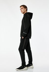 Czarna bluza z kapturem z przednią kieszenią, połączona z czarnymi spodniami z dwoma równoległymi szarymi paskami. Model ma na sobie czarne trampki. Prosty, sportowy design.