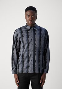 A camisa apresenta um tecido cinza com um padrão de ondas pretas, fecho de botões à frente, colarinho pontudo e um único bolso no peito. Mangas longas.
