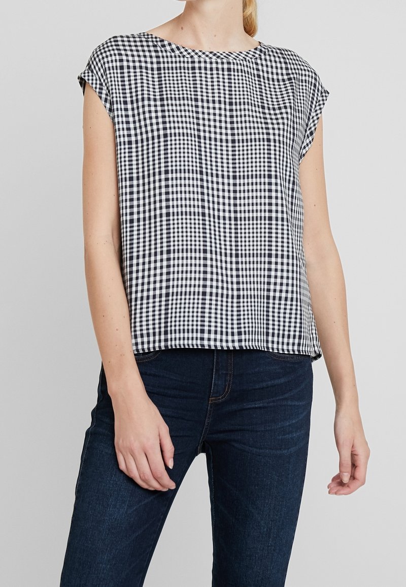 Žena v bezrukávovém topu s černo-bílým kostičkovaným vzorem a tmavě modrých džínách stojící na jednobarevném pozadí.