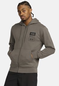 Grijze ritshoodie van zachte stof met een kangoeroezak, featuring zwarte grafische prints en een capuchon met trekkoord.