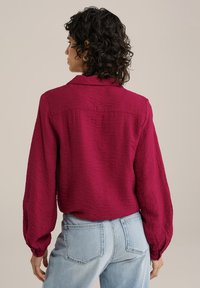 Bordo majica dugih rukava od teksturirane tkanine, s ovratnikom, slobodnim krojem i blago napućenim rukavima, u kombinaciji s svijetloplavim farmerkama.