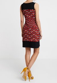 Robe longueur genou présentant un motif géométrique rouge, noir et blanc, avec un haut noir uni et une taille accentuée. Assortie avec des escarpins jaunes.