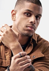 Portrett av en person som har på seg en mørk skjorte og en brun semsket skinnjakke, som holder et håndledd prydet med en metallring og et perlesmykke.