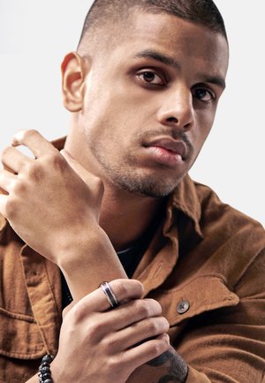 Portrait of a person wearing a dark shirt and brown suede jacket, holding a wrist adorned with a metallic ring and beaded bracelet.