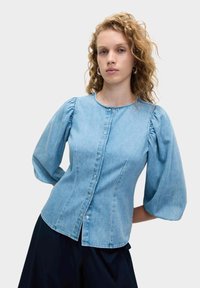 Blusa di denim azzurro chiaro con maniche a palloncino, scollatura rotonda e chiusura con bottoni. Presenta cuciture sagomate e un orlo curvo.