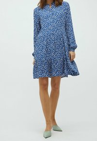 Blaue Kleid mit weißen Blattmustern, langen Ärmeln, knöpfbarem Kragen, geschichteter Rock und einer entspannten Passform. Ein Paar hellgrüne spitze Ballerinas.