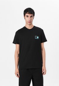 T-shirt en coton noir avec un petit logo multicolore sur la poitrine gauche, col rond, manches courtes, associé à un pantalon noir.