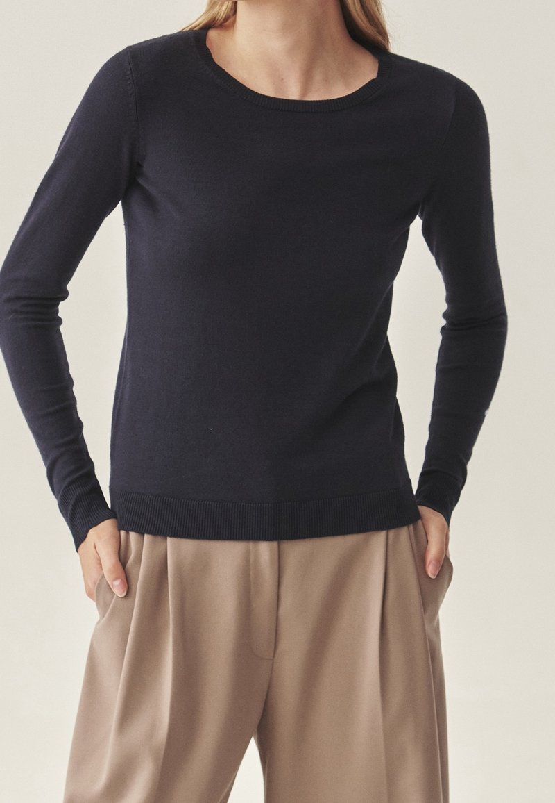 Personne portant un pull noir ajusté à manches longues et un pantalon beige ample, les mains dans les poches, sur un fond neutre.