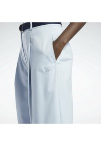 Personne portant un pantalon large bleu clair avec la main dans la poche et un logo blanc Reebok sur la cuisse.
