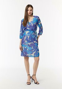Przezroczysta sukienka kopertowa w odcieniach niebieskiego z abstrakcyjnymi wzorami, z rękawami do trzech czwartej i krawatem w talii. Noszona z czarnymi sandałkami na cienkich obcasach.