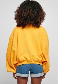 Sudadera amarilla de gran tamaño con mangas abullonadas y dobladillo acanalado, combinada con pantalones cortos vaqueros con puños enrollados y bolsillos traseros.