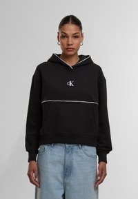 Schwarzer Baumwollhoodie mit weißem Rand, ausgestattet mit einer Vordertasche und einem kleinen gestickten Logo. Kombiniert mit hellblauen Jeans.