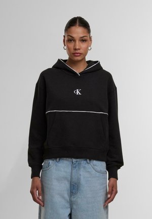 Svart bomullshettegenser med hvit kant, med en lomme foran og en liten brodert logo. Stylet med lyseblå denimjeans.