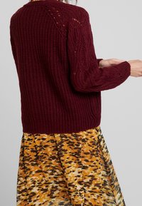 Burgundy knitted sweater with a textured pattern and ribbed hem, paired with a yellow and brown patterned skirt featuring a flared design.