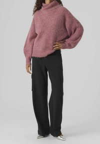 Übergroßer, rosa Strickpullover mit hohem Kragen, kombiniert mit schwarzen, weit geschnittenen Cargo-Hosen und silbernen spitzen Schuhen. Weicher, strukturierter Stoff.