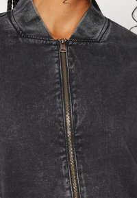 Veste noire à fermeture éclair, en tissu texturé, avec une fermeture éclair dorée contrastante et un col côtelé. Gros plan sur la partie supérieure.