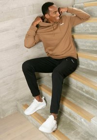 Brauner Kapuzenpullover, schwarze Jogginghose und weiße Sneaker. Sitzt auf Stufen mit Beton- und Holzoberfläche, entspannte Haltung.