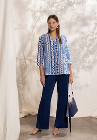 Blusa azul con estampado, cuello en V y mangas tres cuartos, combinada con pantalones anchos azules marinos; la modelo sostiene un bolso azul.