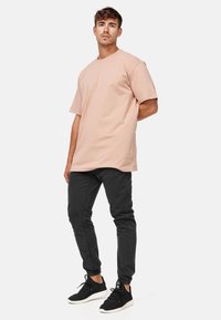 Jeune homme debout portant un t-shirt beige oversize, un jogger noir ajusté et des baskets noires, sur un fond clair uni.