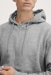 Grå zip-up hettegenser laget av mykt stoff. Har hette med snøring og sølvfargede glidelåsdetaljer. Komfortabel passform med lange ermer.