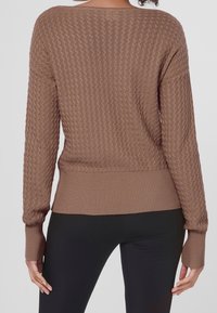 Brązowy sweter z dzianiny o fakturowanym wzorze, szerokim dekolcie oraz ściągaczami przy krawędziach i rękawach. Zestawiony z czarnymi legginsami, prezentując casualowy styl.