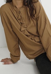 Blusa marrone con scollatura rotonda, chiusura con bottoni e dettagli smerlati lungo la parte anteriore. Maniche lunghe con polsini elasticizzati. Tessuto morbido.