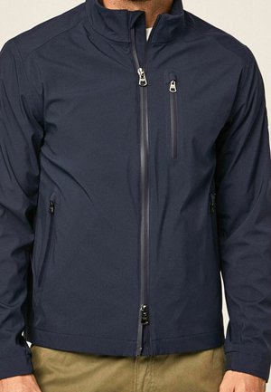 Mann trägt eine marineblaue Jacke mit Reißverschluss und hohem Kragen sowie Reißverschlusstaschen an Brust und Händen, kombiniert mit khakifarbenen Hosen.