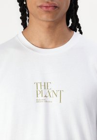 Tricou alb din bumbac cu text imprimat aurit: "PLANTA," cu subtitlul "Colecția Herbarium Toamnă." Design cu guler rotund.