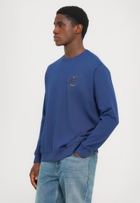 Sudadera de color azul marino con puños y dobladillo acanalados, que presenta un logo bordado en el pecho. Tejido suave, diseño clásico de cuello redondo.